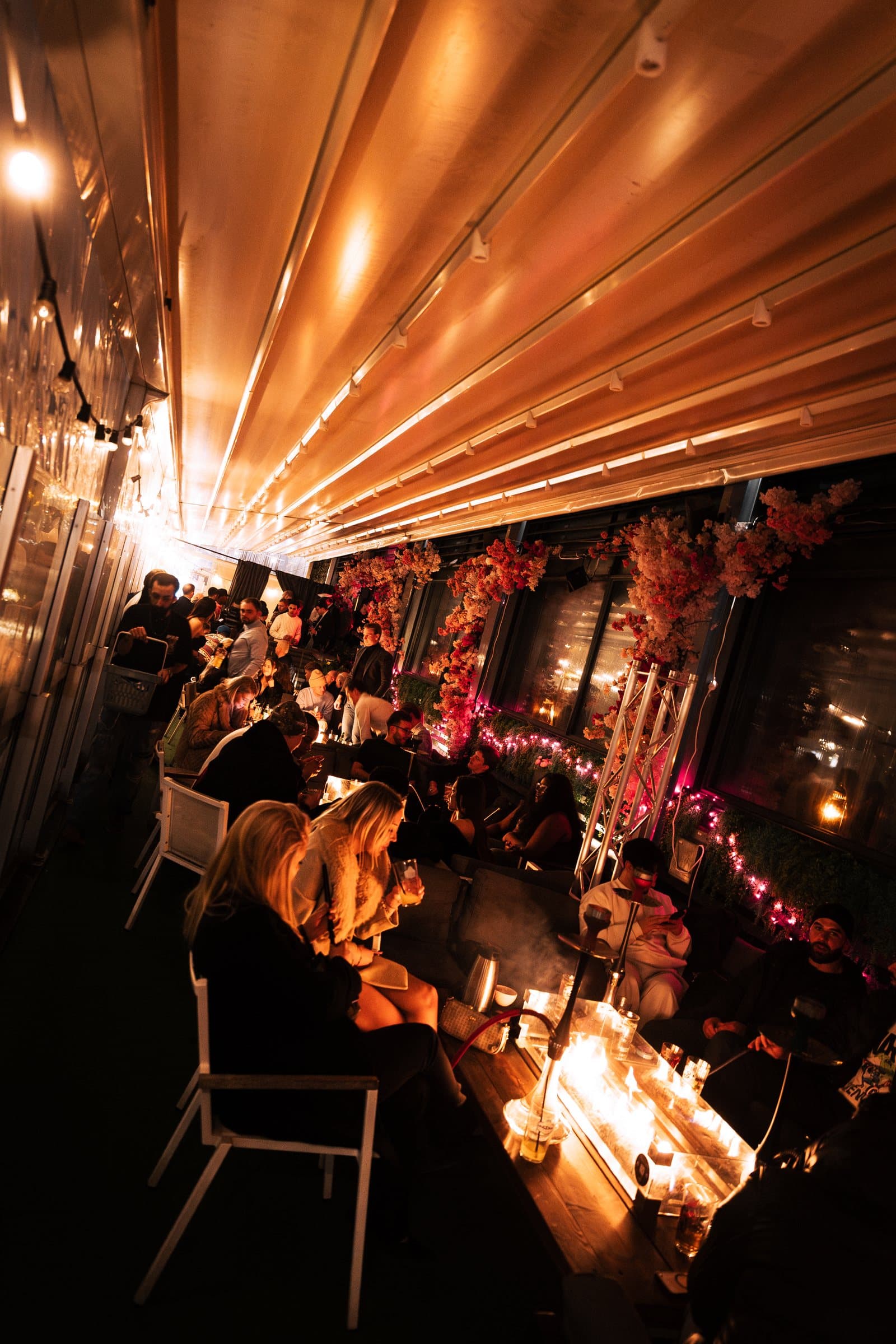 Guests on the heated glass terrace among blossoms and warm lights, water pipes on the fire tables
